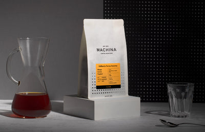 A bag of Machina coffee on a grey block beside a glass, spoon and pourer.